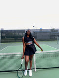 Woman on a tennis court in a black tennis outfit holding a racket and a ball