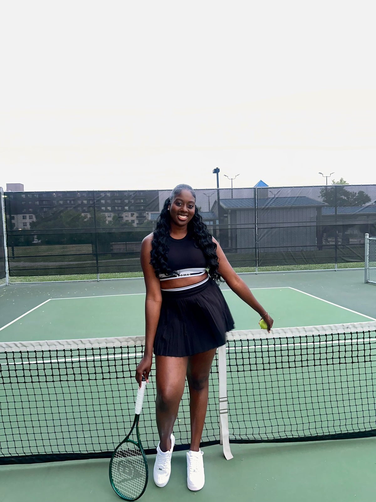 Woman on a tennis court in a black tennis outfit holding a racket and a ball