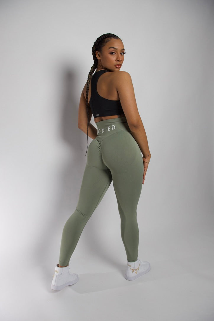 A person wearing green full-length leggings with a unique scrunch seam detail, and a black sports bra, standing against a plain background.